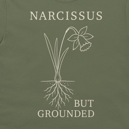 Close-up of an olive green t-shirt featuring a white line art graphic of a daffodil with its roots exposed, accompanied by the text 'NARCISSUS BUT GROUNDED' in a matching off-white serif font.