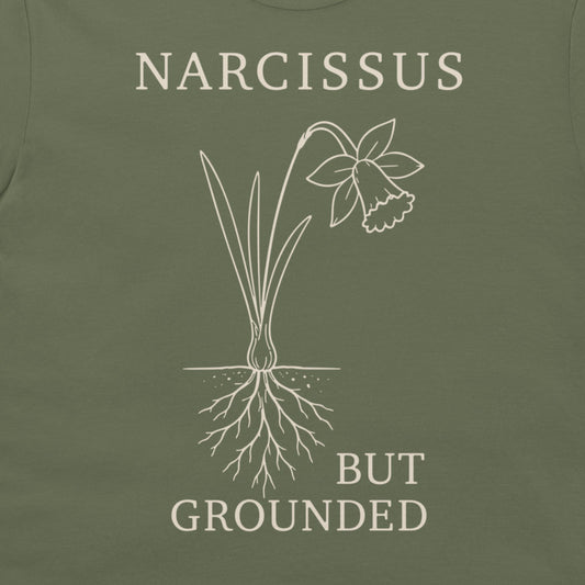 Close-up of an olive green t-shirt featuring a white line art graphic of a daffodil with its roots exposed, accompanied by the text 'NARCISSUS BUT GROUNDED' in a matching off-white serif font.