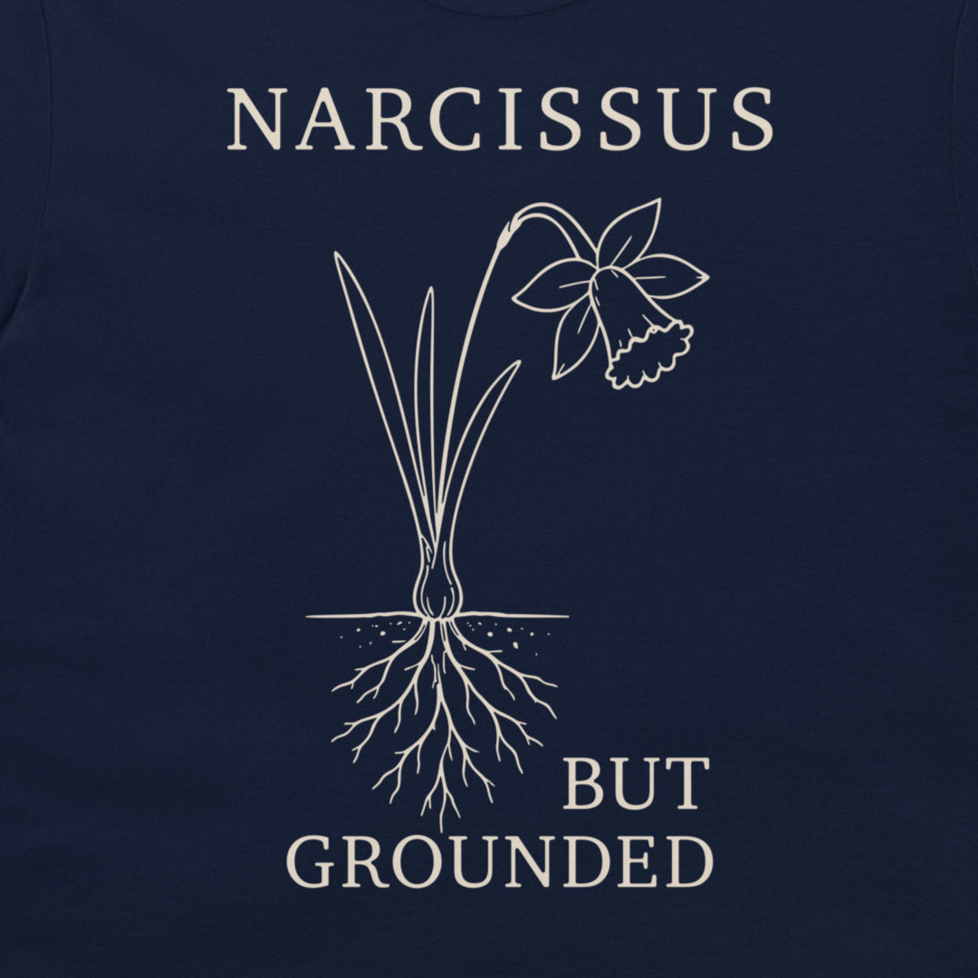 A close-up of a navy blue t-shirt, featuring a white graphic print of a daffodil with its roots exposed and the text 'NARCISSUS BUT GROUNDED' in a serif font.