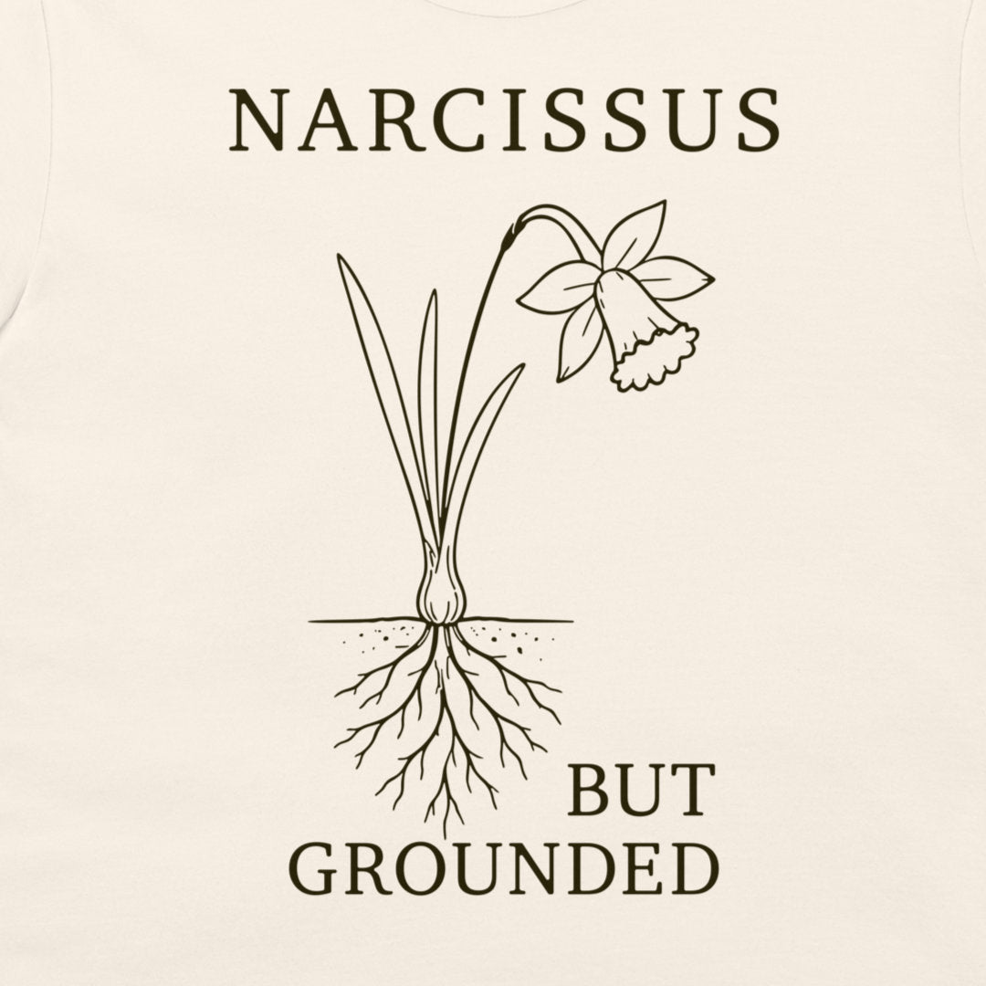 A close-up of a cream-colored t-shirt, featuring a dark brown graphic print of a daffodil with its roots exposed, accompanied by the text 'NARCISSUS BUT GROUNDED'.