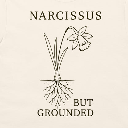 A close-up of a cream-colored t-shirt, featuring a dark brown graphic print of a daffodil with its roots exposed, accompanied by the text 'NARCISSUS BUT GROUNDED'.