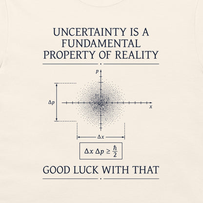 A cream-colored graphic t-shirt featuring white text and a physics diagram. The text reads 'UNCERTAINTY IS A FUNDAMENTAL PROPERTY OF REALITY' at the top and 'GOOD LUCK WITH THAT' at the bottom, surrounding a central uncertainty principle diagram.