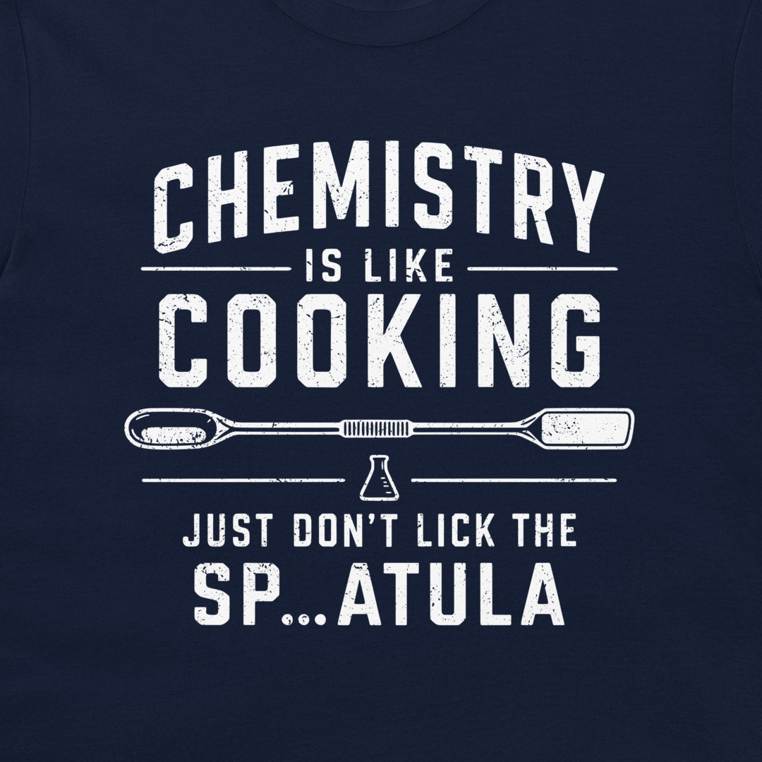 A close-up of a navy blue shirt featuring a distressed white graphic print that reads 'CHEMISTRY IS LIKE COOKING' with a beaker and spatula design, and 'JUST DON'T LICK THE SP...ATULA'.