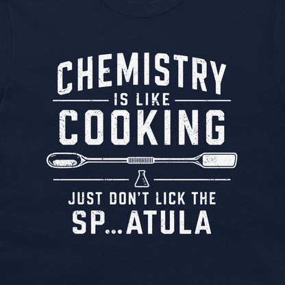A close-up of a navy blue shirt featuring a distressed white graphic print that reads 'CHEMISTRY IS LIKE COOKING' with a beaker and spatula design, and 'JUST DON'T LICK THE SP...ATULA'.