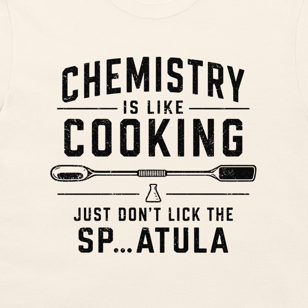 A close-up of a light cream-colored t-shirt featuring a black distressed graphic print that reads 'CHEMISTRY IS LIKE COOKING' with a spatula illustration and 'JUST DON'T LICK THE SP...ATULA'.