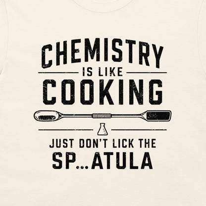 A close-up of a light cream-colored t-shirt featuring a black distressed graphic print that reads 'CHEMISTRY IS LIKE COOKING' with a spatula illustration and 'JUST DON'T LICK THE SP...ATULA'.