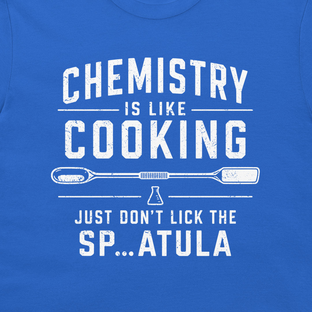 A close-up of a blue t-shirt featuring a white distressed graphic that reads 'CHEMISTRY IS LIKE COOKING' with a spatula and beaker illustration, followed by 'JUST DON'T LICK THE SP...ATULA'.