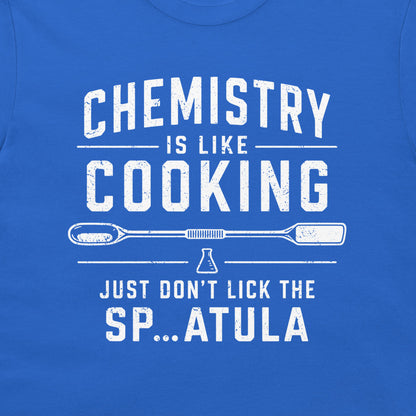 A close-up of a blue t-shirt featuring a white distressed graphic that reads 'CHEMISTRY IS LIKE COOKING' with a spatula and beaker illustration, followed by 'JUST DON'T LICK THE SP...ATULA'.