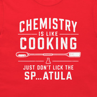 A close-up of a bright red t-shirt, featuring a distressed white graphic print that reads 'CHEMISTRY IS LIKE COOKING' and 'JUST DON'T LICK THE SP...ATULA'.