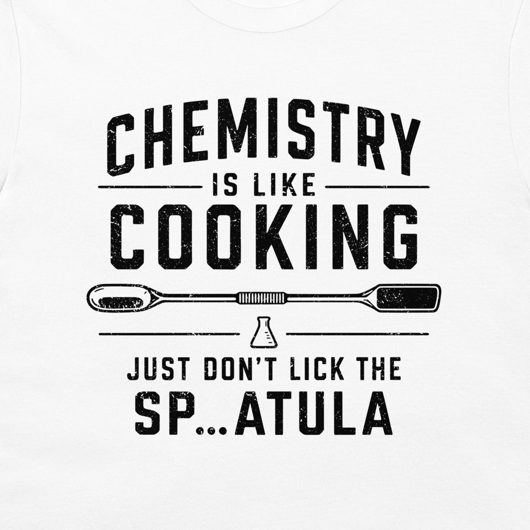 A white t-shirt featuring a black distressed graphic print that reads 'CHEMISTRY IS LIKE COOKING' with a spatula and beaker illustration, followed by 'JUST DON'T LICK THE SP...ATULA'.