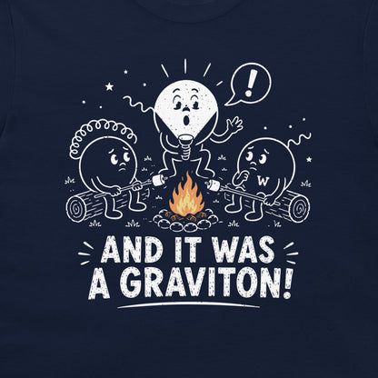 A close-up of a navy blue t-shirt featuring a white graphic of three cartoon particle characters roasting marshmallows over a campfire, with the text 'AND IT WAS A GRAVITON!' printed below.