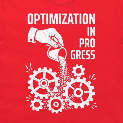 A close-up of a red garment, likely a t-shirt, featuring a white distressed graphic print that reads 'OPTIMIZATION IN PROGRESS' above a hand pouring sand into gears.