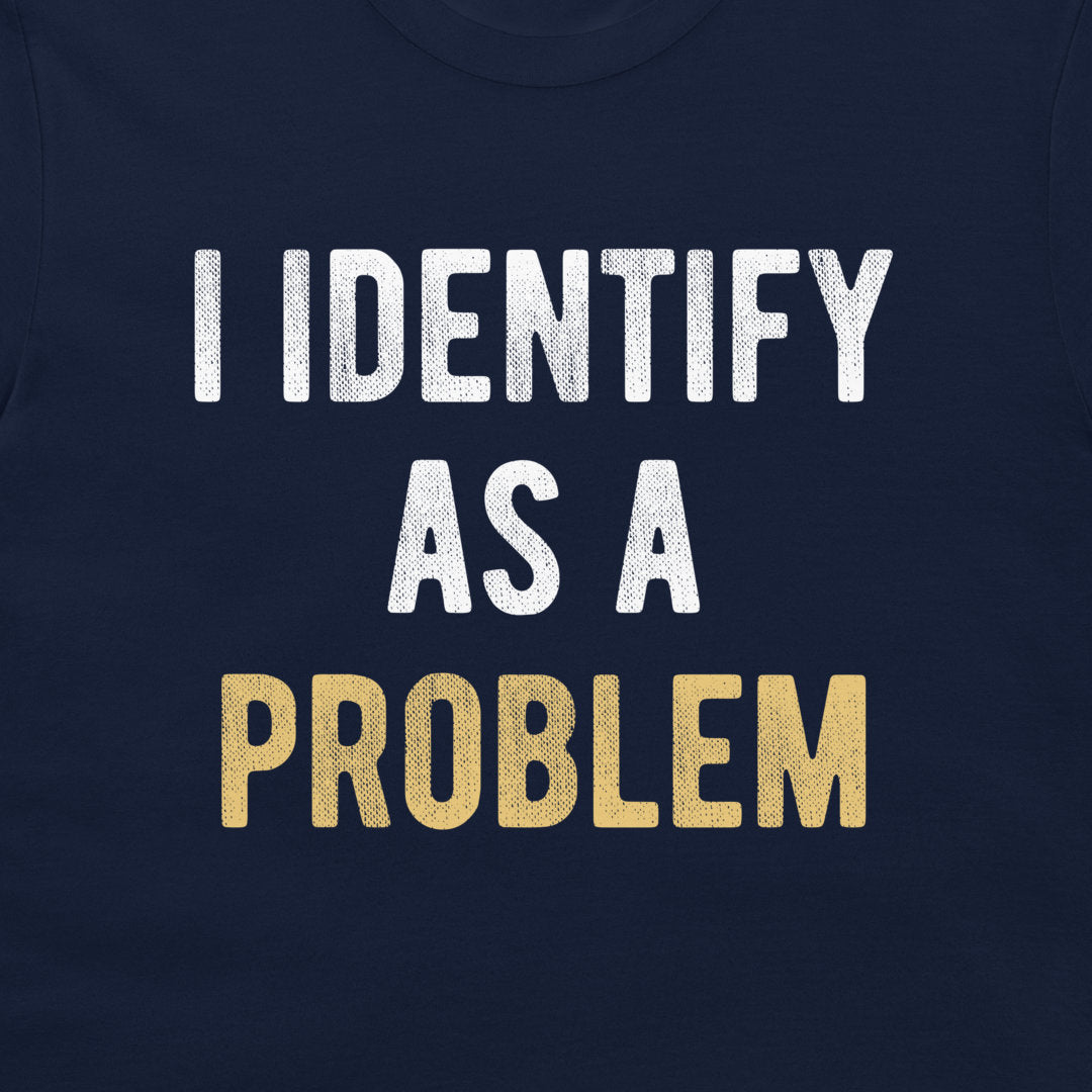 A close-up of a navy blue t-shirt featuring a distressed text graphic that reads 'I IDENTIFY AS A PROBLEM' in white and gold lettering.