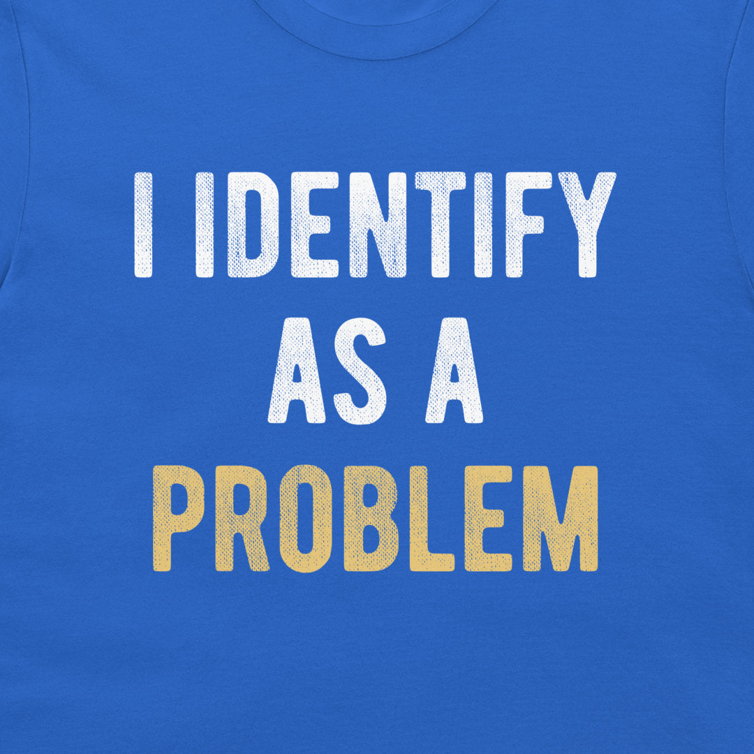 A close-up of a blue T-shirt with the phrase 'I IDENTIFY AS A PROBLEM' printed across the chest in large distressed letters. The first three lines are white, and the bottom line is yellow.