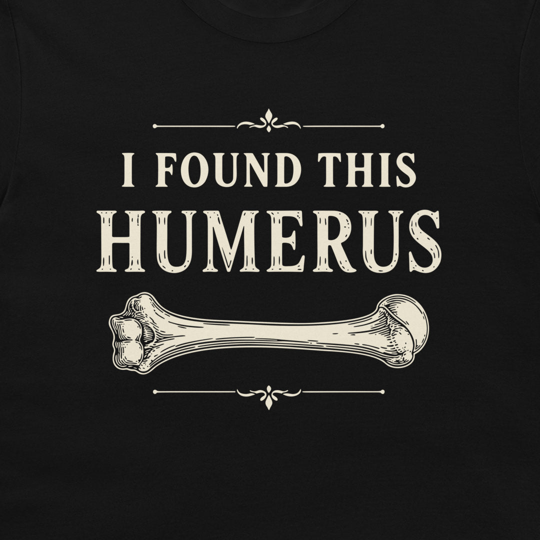 A close-up of a black garment featuring a distressed cream-colored graphic print that reads 'I FOUND THIS HUMERUS' above and below a detailed illustration of a bone.