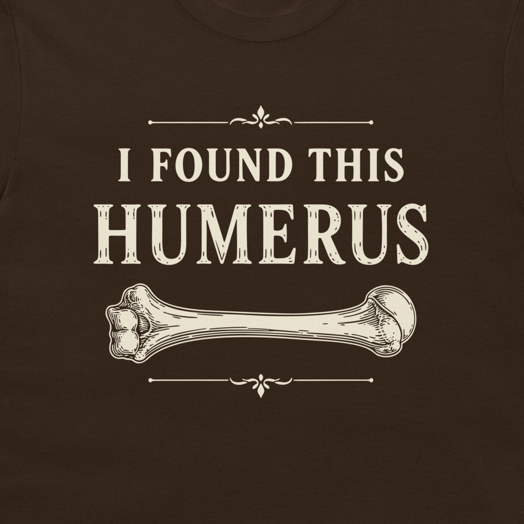 A close-up of a dark brown t-shirt featuring a vintage-style cream graphic that reads 'I FOUND THIS HUMERUS' with an illustration of a bone.
