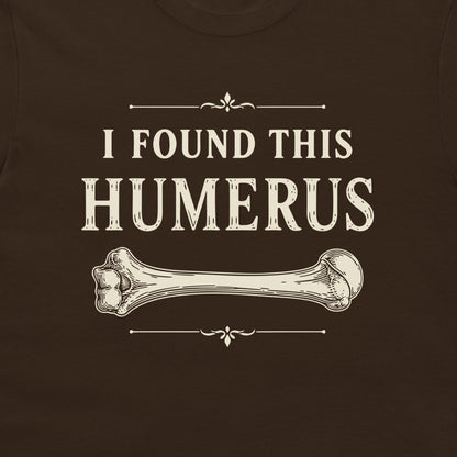 A close-up of a dark brown t-shirt featuring a vintage-style cream graphic that reads 'I FOUND THIS HUMERUS' with an illustration of a bone.