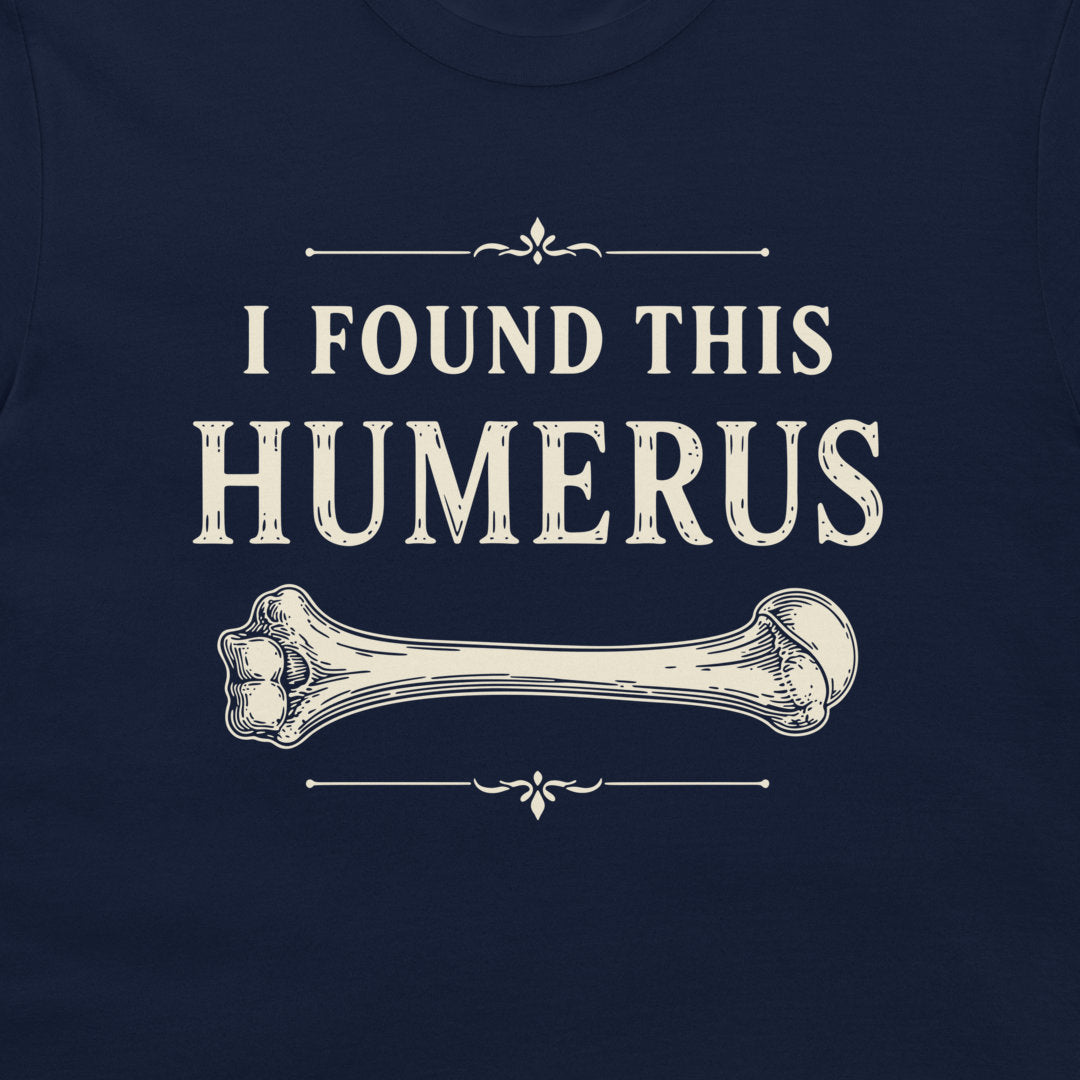 A close-up of a navy blue garment, likely a t-shirt, featuring a vintage-style cream graphic that reads 'I FOUND THIS HUMERUS' above an illustration of a bone.