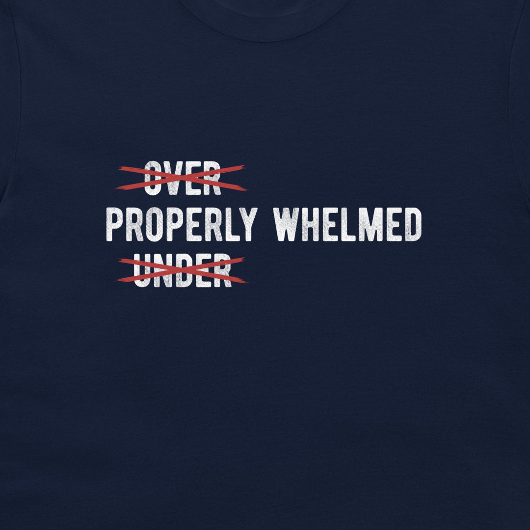 A close-up of a navy blue t-shirt featuring a graphic print with the words 'OVER' and 'UNDER' in white, both crossed out with red scribbles, with 'PROPERLY WHELMED' printed in white between them.