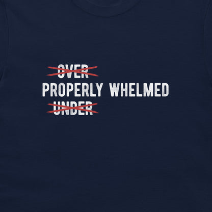A close-up of a navy blue t-shirt featuring a graphic print with the words 'OVER' and 'UNDER' in white, both crossed out with red scribbles, with 'PROPERLY WHELMED' printed in white between them.