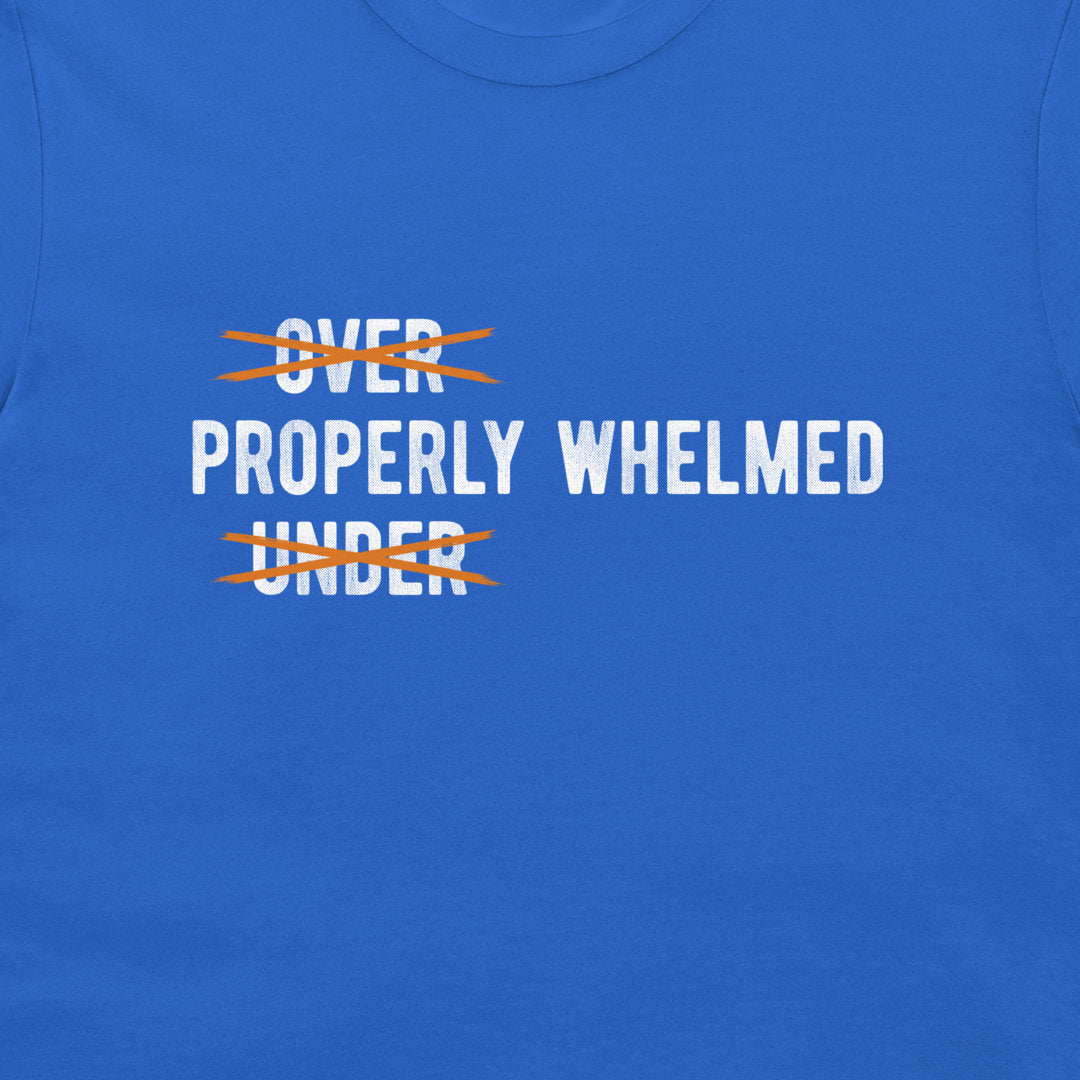 A close-up of a royal blue t-shirt featuring a graphic print. The text reads 'PROPERLY WHELMED' in white capital letters, with 'OVER' and 'UNDER' crossed out in orange scribbles.