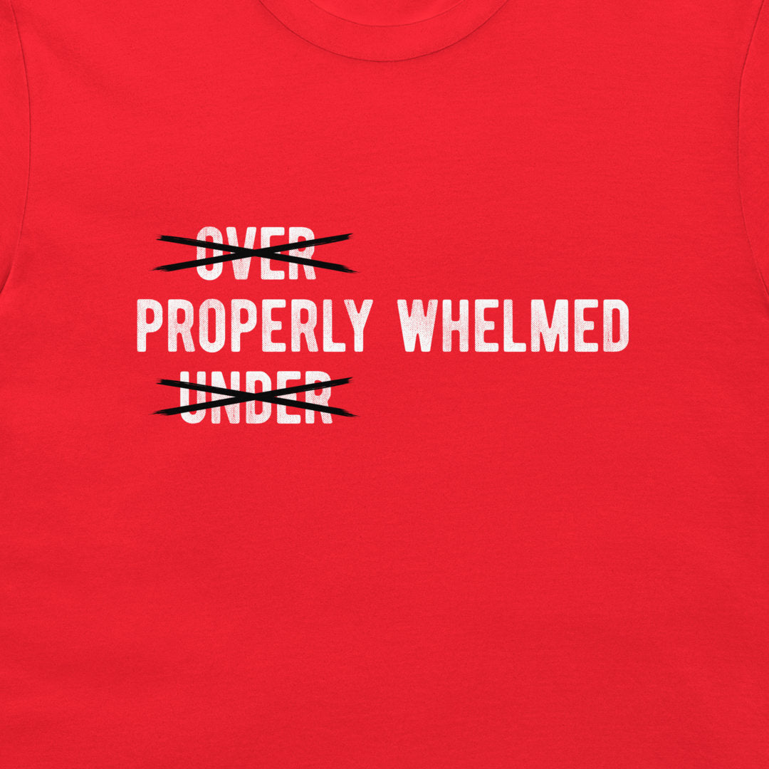 A close-up of a bright red t-shirt featuring a graphic print. The design displays the words 'OVER PROPERLY WHELMED UNDER' in white capital letters, with 'OVER' and 'UNDER' crossed out in black scribbles.