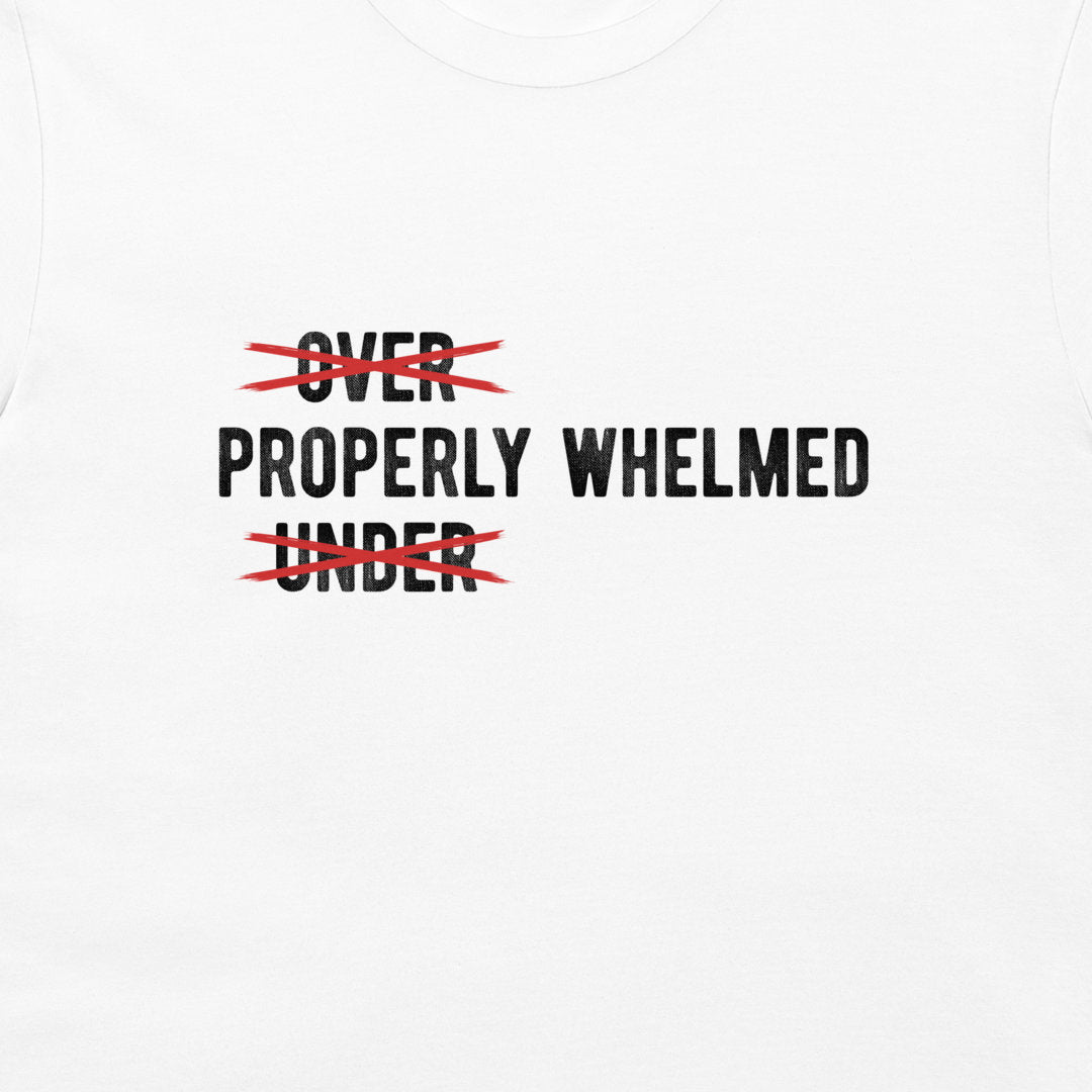 White t-shirt featuring a graphic print with the words 'OVER' and 'UNDER' in black text, both crossed out with red scribbles, leaving 'PROPERLY WHELMED' printed in the center.